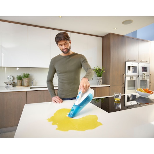 A person using a BLACK+DECKER handheld vacuum cleaner (model WDA315J) to clean a liquid spill on a kitchen countertop. The kitchen is modern with white and wooden cabinets, plants, and a bowl of fruit visible.