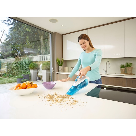A person is using a BLUE BLACK+DECKER handheld vacuum cleaner to clean crumbs from a white kitchen countertop. There is a bowl of fruit and a purple bowl on the counter, and the kitchen has large windows showing a garden outside.