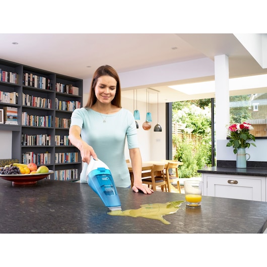A person using a BLACK+DECKER handheld vacuum cleaner (model WDC115WA) to clean up a spilled liquid on a kitchen countertop. The background shows a modern kitchen and dining area with bookshelves, fruit, and flowers.