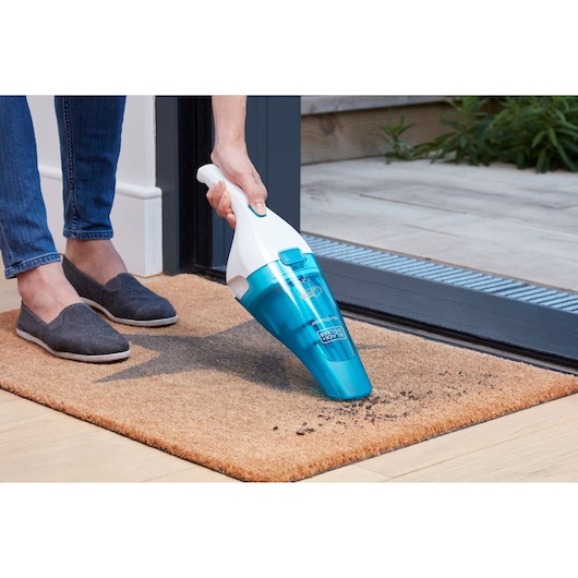 A person uses a BLUE BLACK+DECKER handheld vacuum cleaner to clean dirt from a doormat near a doorway.