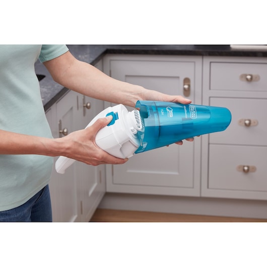 Person holding and opening a BLUE BLACK+DECKER handheld vacuum cleaner over a kitchen counter, demonstrating how to remove the dust container.