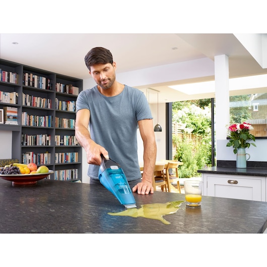 A person using a BLUE BLACK+DECKER handheld vacuum cleaner (model WDC215WA) to clean spilled juice on a kitchen countertop, with a glass of juice and a bowl of fruit nearby.